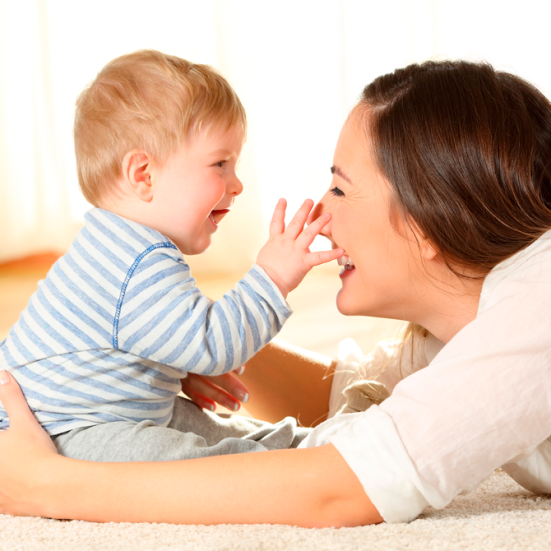 Baby sidder på gulvet og rækker ud mod kvindes ansigt. Kvinde ligger på maven med armene omkring baby. 