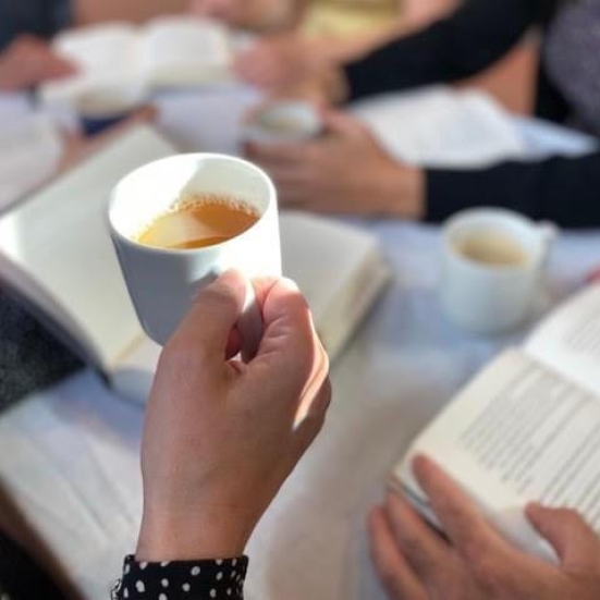 Billede af hånd, der holder en kop med kaffe. Foran et bord med hænder og bøger. 