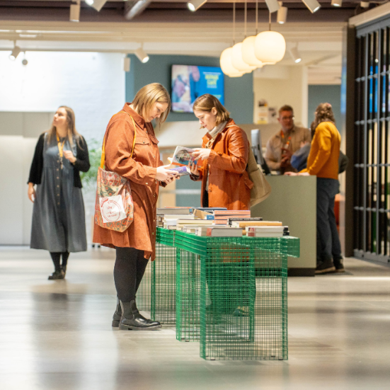 Billede af to biblioteksbrugere ved et grønt trådbord. De kigger på bøger. Bibliotekspersonale i baggrunden. 
