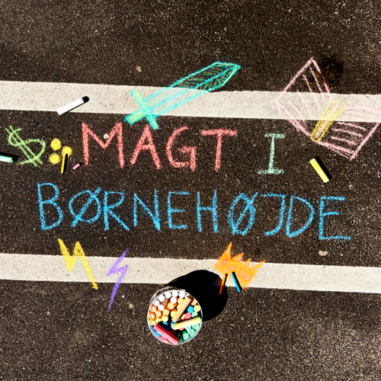 Sort asfalt med kridttegning. Åben bog, sværd, lyn og dollartegn. Med teksten magt i børnehøjde