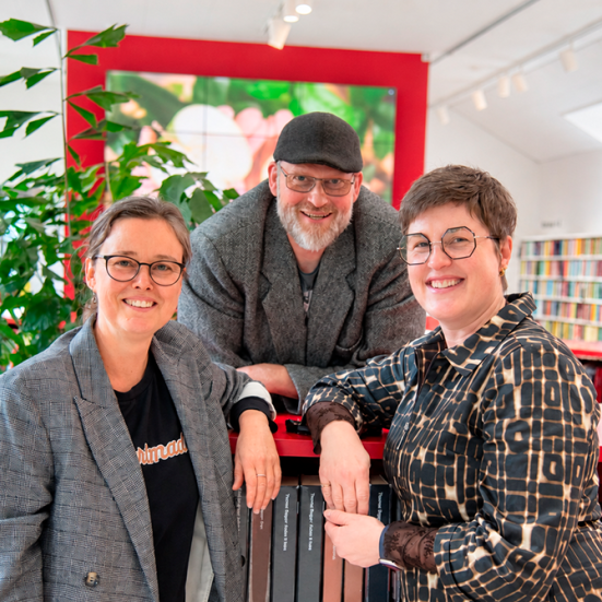 Tre mennesker står samlet i et lyst biblioteksrum.