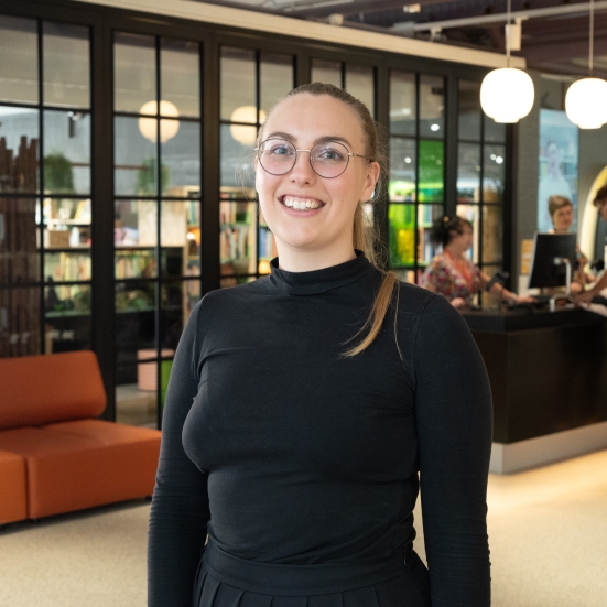 Kvinde med mørk trøje og briller på et bibliotek