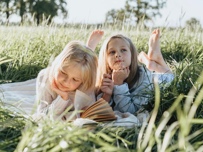 To piger ligger på maven i en græsmark og læser mens det er sommer