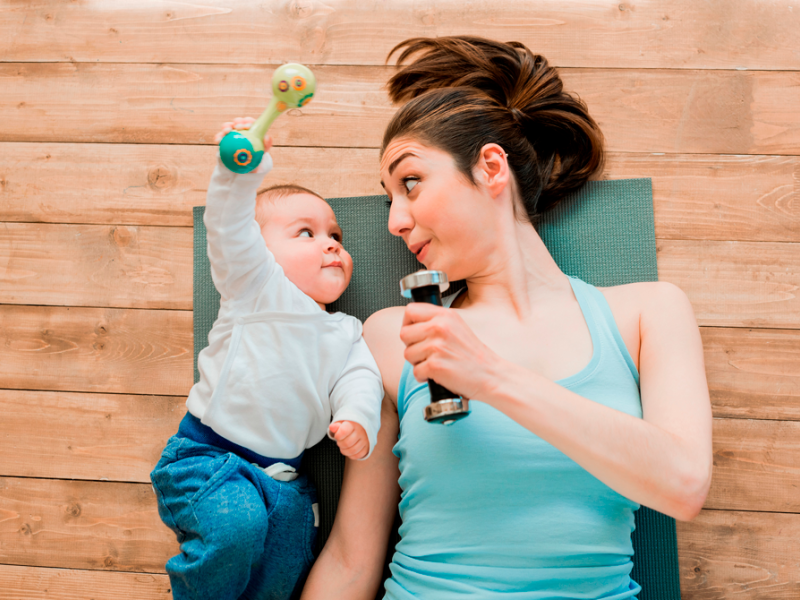 Baby og kvinde ligger på jorden på en yogamåtte med træningsudstyr. De kigger på hinanden