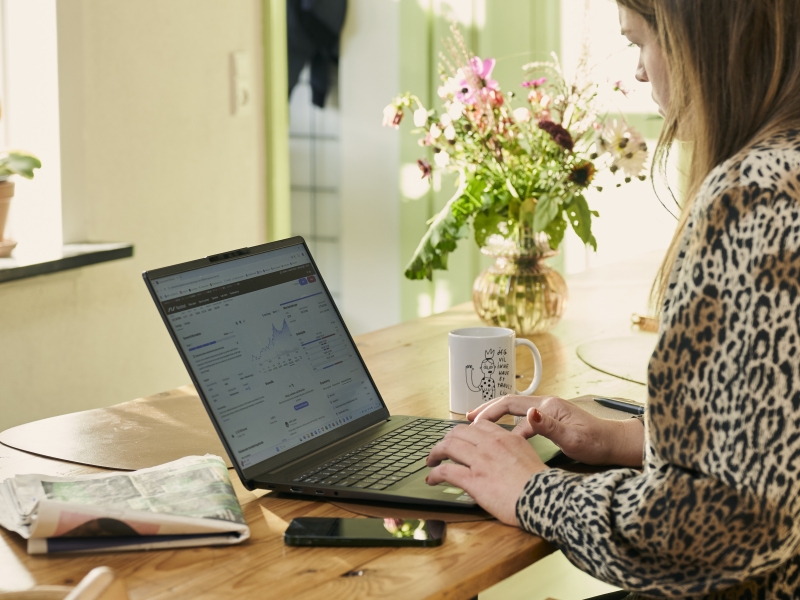 kvinde set fra siden, arbejder på pc, i baggrunden kaffekop og blomsterbuket