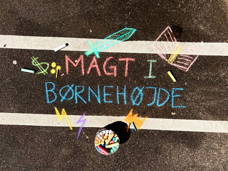 Sort asfalt med kridttegning. Åben bog, sværd, lyn og dollartegn. Med teksten magt i børnehøjde