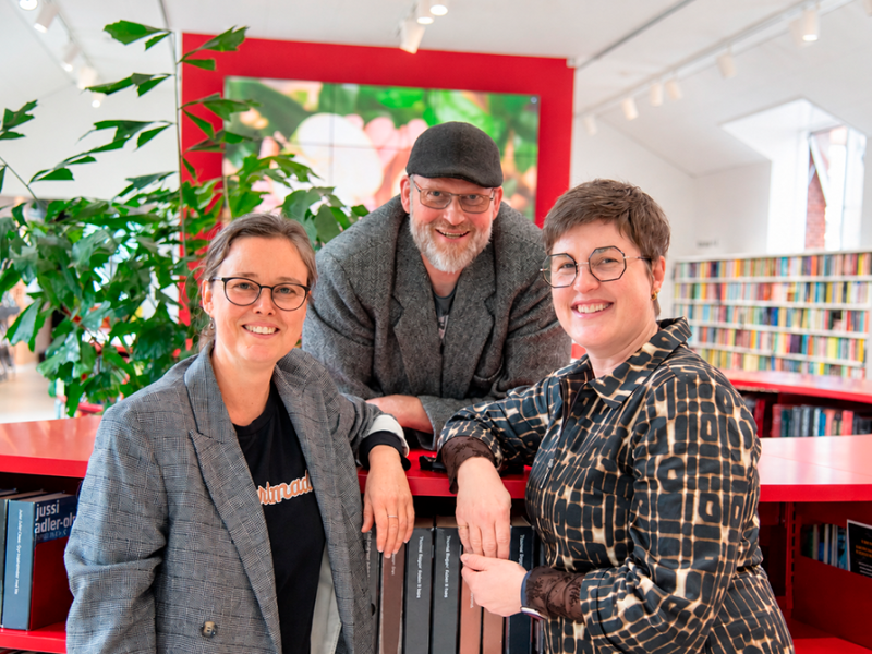Tre mennesker står samlet i et lyst biblioteksrum.