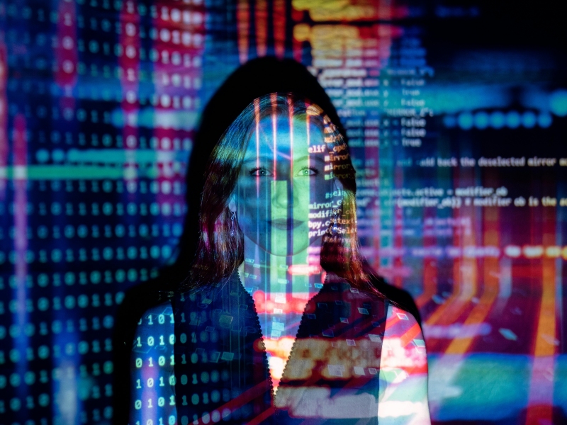 kvinde står i baggrunden mens flere billeder med data, talkolonner og programmeringskode i mange farver bliver projekteret på hende og væggen bagved