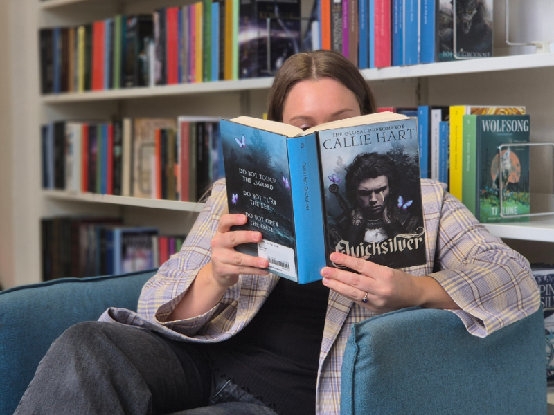 Kvinde i blå lænestol foran bogreol, som læser Quicksilver-bogen. 
