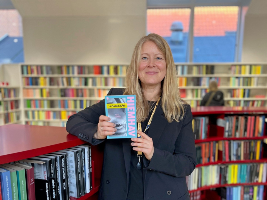 Formidler Trine i biblioteksrummet, lænet op ad en rød reol med bogen "Hjemhav" i hænderne