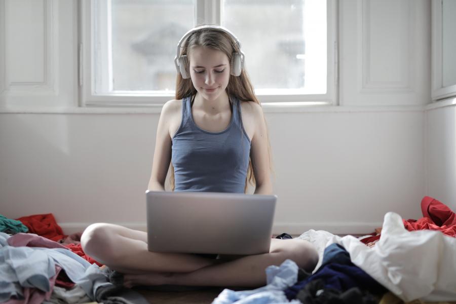 teenager med hovedtelefoner ser på bærbar pc, mens der er spredt vasketøj på gulvet rundt om personen